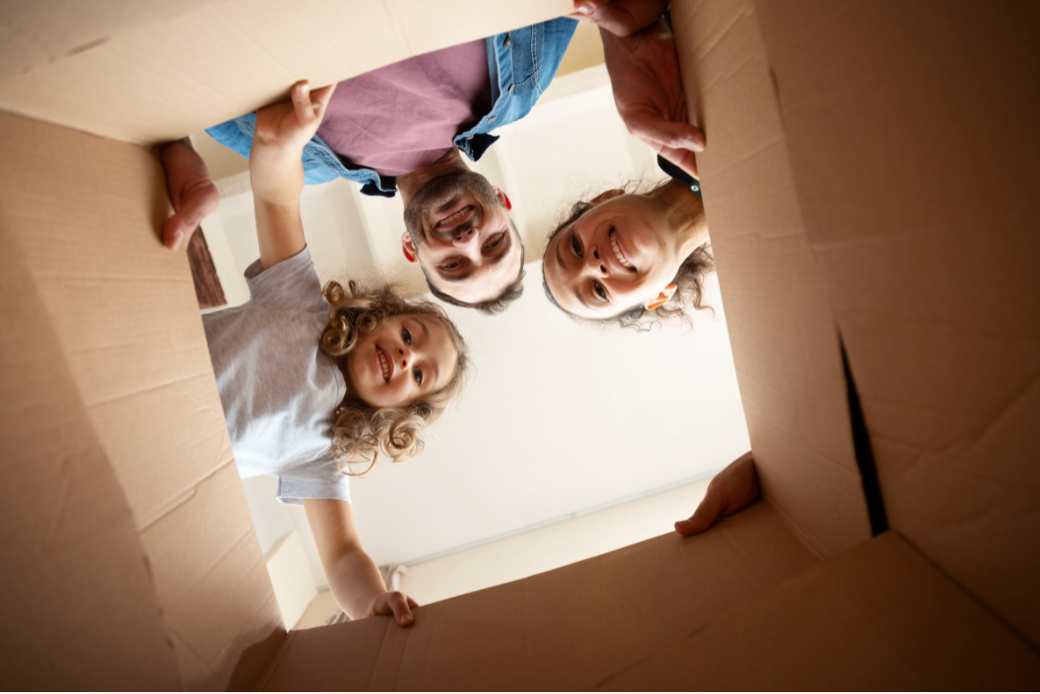 Photo prise par le dessous d'un carton de déménagement : au dessus, on voit les visages souriants d'une famille