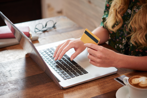 Photo représentant une femme dont on ne voit pas le visage, tapant sur le clavier de l'ordinateur avec l'une de ses mains et avec sa carte bleue dans l'autre.