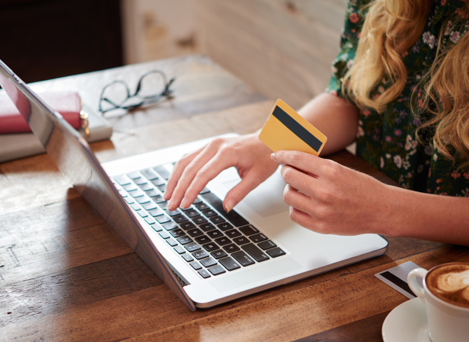 Photo représentant une femme dont on ne voit pas le visage, tapant sur le clavier de l'ordinateur avec l'une de ses mains et avec sa carte bleue dans l'autre.