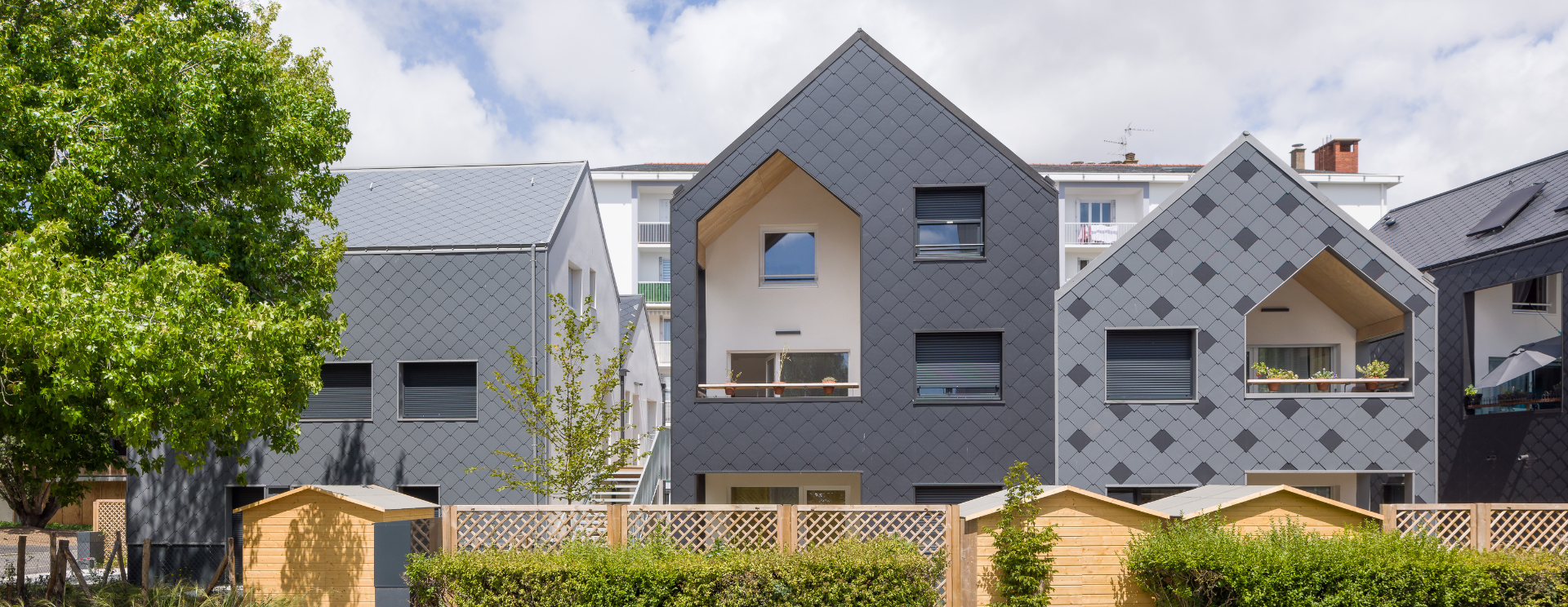 Photographie de la résidenceLes loges à Saint-Nazaire. Un résidence semi collective avec un étage, à l'esprit de maison avec des murs recouverts d'ardoise, dans les tons gris. Au premier plan, les clôtures en bois et la haie.