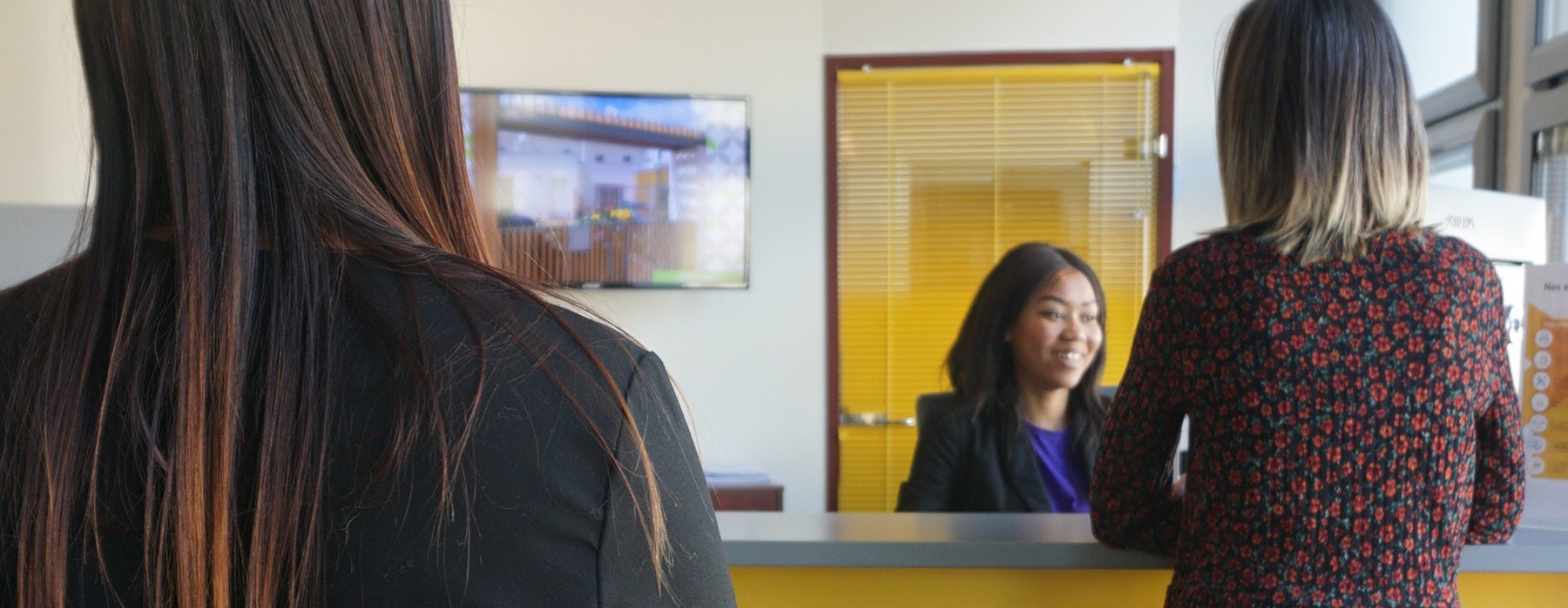 Photo d'une chargée de clientèle de face assise devant le comptoir d'accueil, deux clients de dos lui font face.