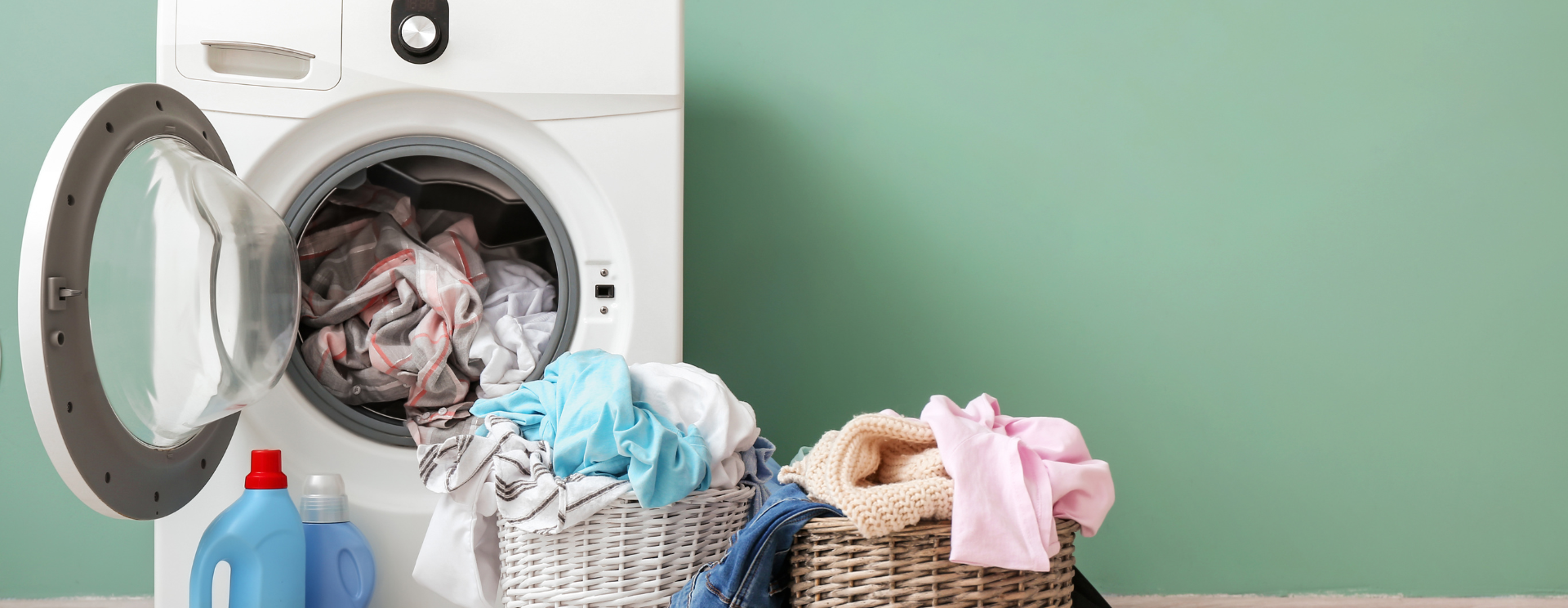 Photo d'une machine à laver devant un mur vert, avec deux petits paniers à linge rempli juste devant.