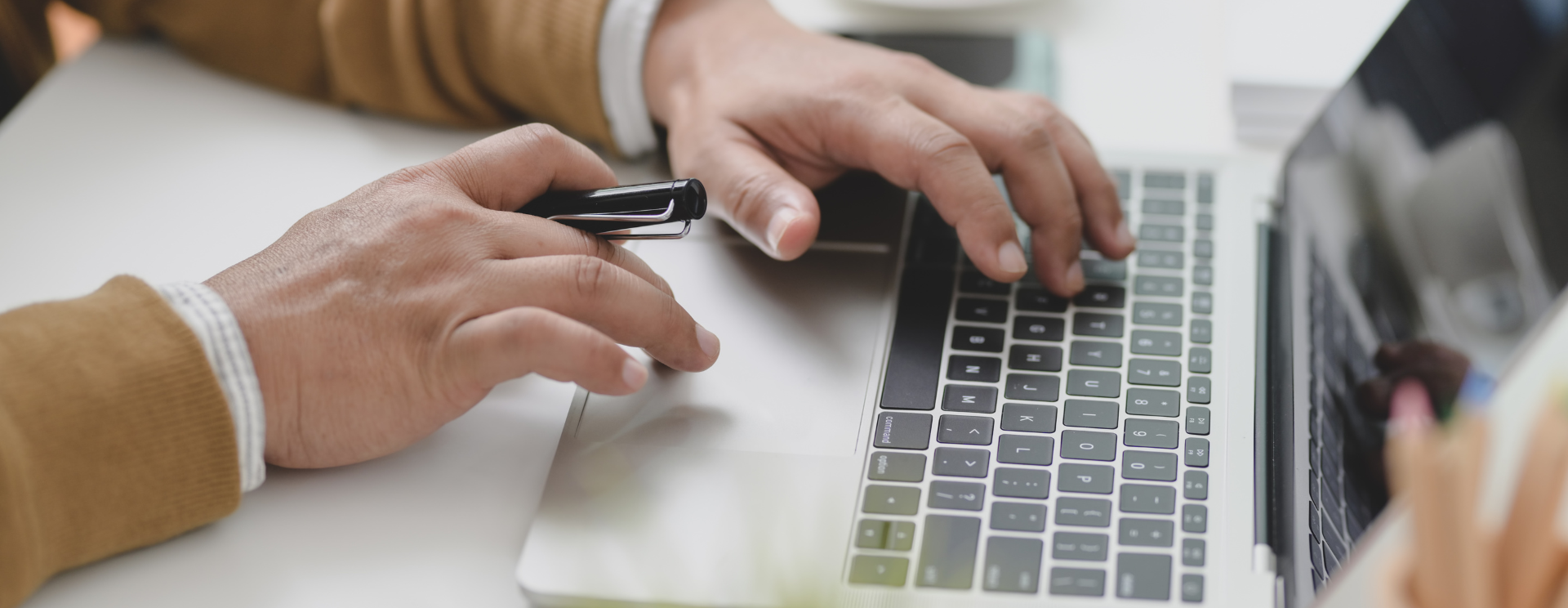 Photo en gros plan sur les mains d'un homme qui est en train de taper à l'ordinateur, il tient un crayon à la main.