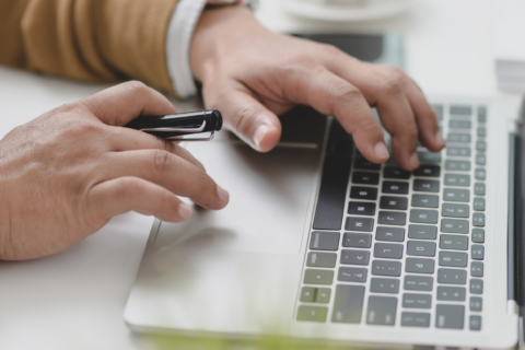 Photo en gros plan sur les mains d'un homme qui est en train de taper à l'ordinateur, il tient un crayon à la main.
